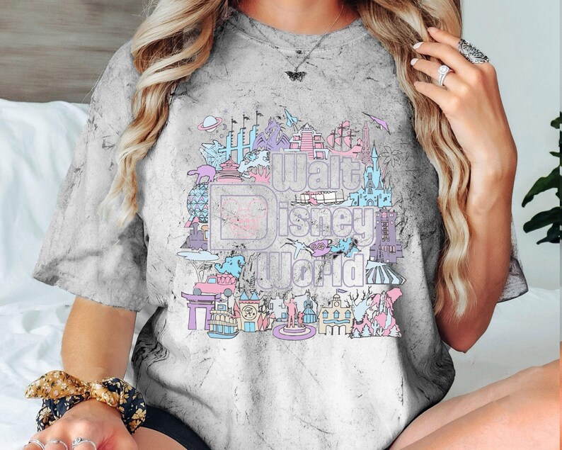 May include: Grey tie-dye t-shirt with a pastel graphic of the Walt Disney World logo and park icons. The design includes castles, rockets, and other attractions in pink, blue, and purple.