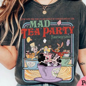 Puede incluir: Camiseta gris oscuro con un gr&aacute;fico colorido de la Mad Tea Party de Fantasyland de Disney. El dise&ntilde;o incluye personajes como el Sombrerero Loco, Alicia y Mickey y Minnie Mouse en tazas de t&eacute;. El texto dice "MAD TEA PARTY" y "FANTASYLAND".