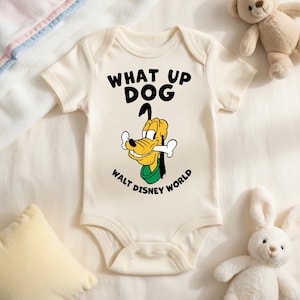 May include: Cream-colored baby onesie with the phrase "WHAT UP DOG" in black letters. Below the text is a cartoon image of Pluto holding a bone, with "WALT DISNEY WORLD" printed below the image.