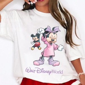 May include: White t-shirt featuring a cartoon Minnie Mouse in a pink dress holding a small Mickey Mouse. The text "Walt Disney World" is printed in purple below the characters. The shirt has short sleeves.