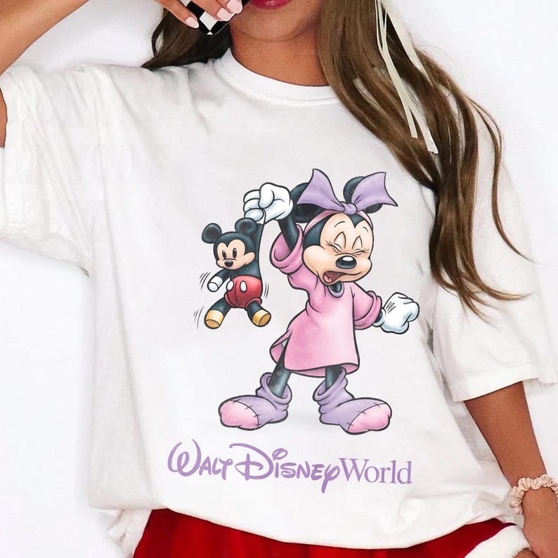 May include: White t-shirt featuring a cartoon Minnie Mouse in a pink dress holding a small Mickey Mouse. The text "Walt Disney World" is printed in purple below the characters. The shirt has short sleeves.