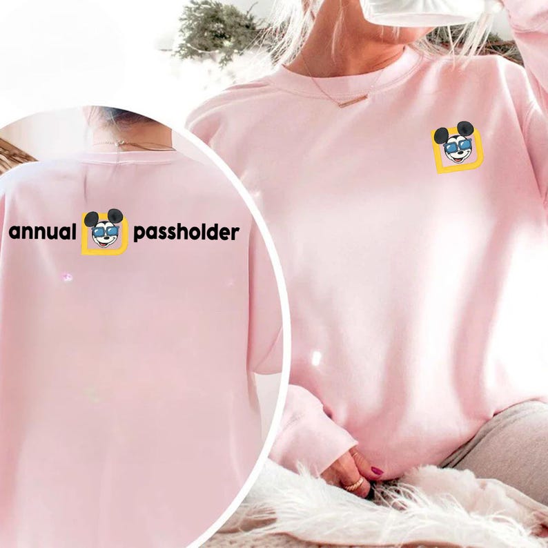Puede incluir: Sudadera rosa claro con un dibujo de Mickey Mouse con gafas de sol en un cuadrado amarillo. El texto "annual passholder" est&aacute; impreso en la espalda. La sudadera es de cuello redondo.
