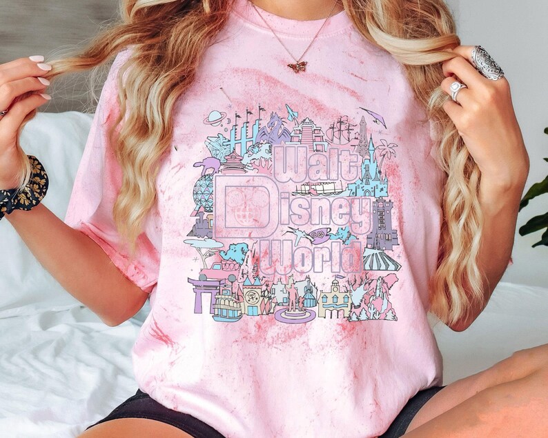 May include: Pink tie-dye t-shirt with a pastel graphic of Walt Disney World landmarks and the park's logo. The design includes illustrations of the castle, Spaceship Earth, and other attractions. The text "Walt Disney World" is prominently displayed.
