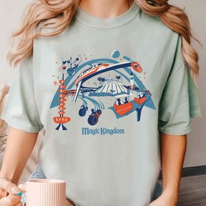 Op de afbeelding: Lichtgroen t-shirt met een kleurrijke illustratie van een pretpark, inclusief een monorail, een raket en andere attracties. De tekst "Magic Kingdom" staat onder de afbeelding en "LAND" staat links.