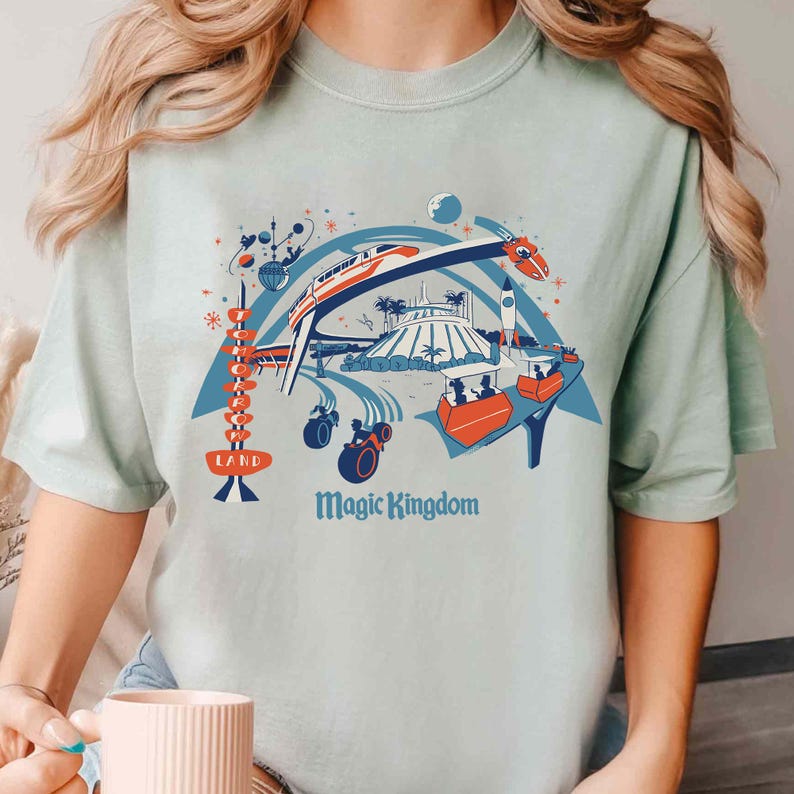 Op de afbeelding: Lichtgroen t-shirt met een kleurrijke illustratie van een pretpark, inclusief een monorail, een raket en andere attracties. De tekst "Magic Kingdom" staat onder de afbeelding en "LAND" staat links.