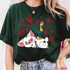 May include: Dark green t-shirt featuring a cartoon illustration of a white pig lying on its back with a blue and green rooster standing on top. Red palm trees and suns are in the background. The shirt has rolled-up sleeves.