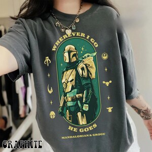 Peut inclure: T-shirt graphite avec un motif du Mandalorien et Grogu. Le design comprend le texte "WHEREVER I GO HE GOES MANDALORIAN & GROGU" en vert et or. Des ic&ocirc;nes sur le th&egrave;me de Star Wars sont dispers&eacute;es autour du motif principal.