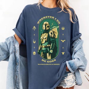 Peut inclure: T-shirt bleu marine avec un motif du Mandalorien et Grogu. Le design comprend le texte "WHEREVER I GO HE GOES" et "MANDALORIAN & GROGU" en vert et or. Le t-shirt a des manches retrouss&eacute;es.