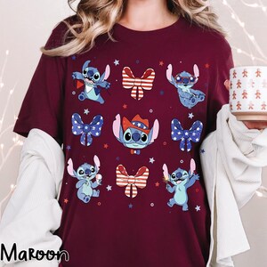 May include: A maroon t-shirt with a pattern of Stitch characters in patriotic poses, surrounded by bow ties and stars. The shirt has a crew neck and short sleeves. The word "Maroon" is printed at the bottom left.