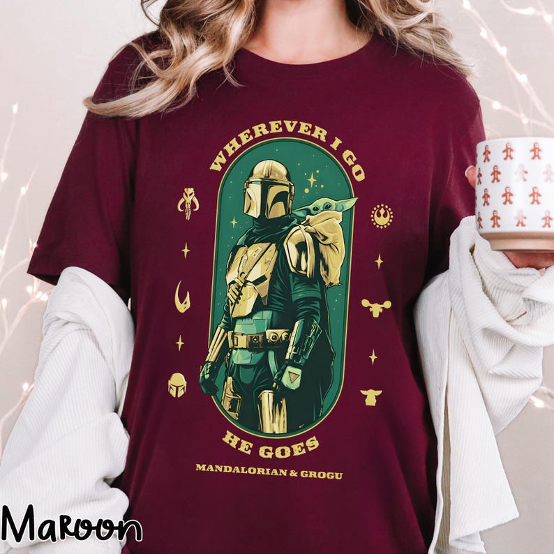 Peut inclure: T-shirt bordeaux avec un motif de The Mandalorian et Grogu. Le design comprend le texte "WHEREVER I GO HE GOES" et "MANDALORIAN & GROGU" en or et blanc. L'illustration est encadr&eacute;e dans une arche verte.