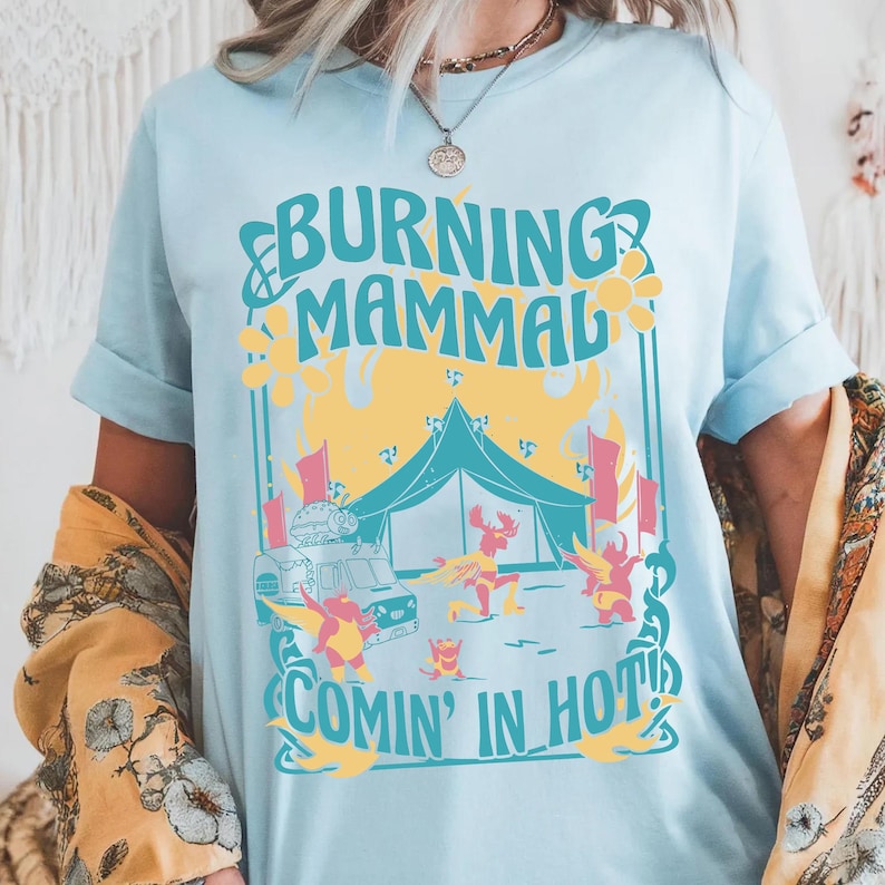 Puede incluir: Camiseta azul claro con un gr&aacute;fico colorido. El dise&ntilde;o presenta el texto "BURNING MAMMAL COMIN' IN HOT!" con ilustraciones de una tienda de campa&ntilde;a, una furgoneta y figuras bailando. La camiseta tiene mangas enrolladas.