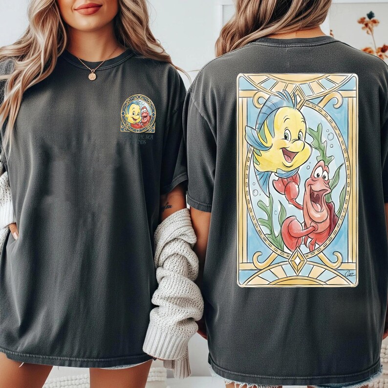 May include: Dark gray t-shirts featuring a colorful illustration of Flounder and Sebastian from The Little Mermaid. The front of the shirt has a smaller circular graphic with the same characters. The back has a larger rectangular design.