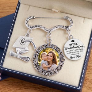 May include: Silver-toned graduation brooch with a photo pendant and charms. The brooch includes a graduation cap charm, a scroll charm, and a round pendant with the text "ON YOUR Graduation Day...Love, Mom."