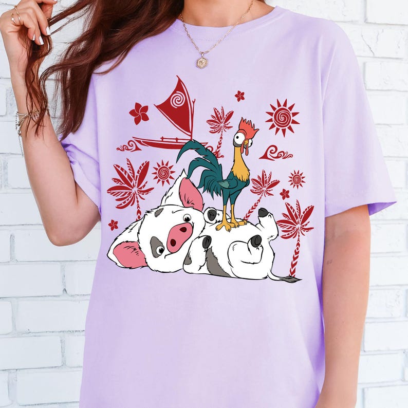 May include: A light purple t-shirt featuring a colorful graphic of cartoon characters. The design includes a pig, a rooster, a boat, and various tropical elements in red, yellow, and green. The shirt is made of a soft material.