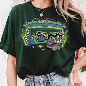 K&ouml;nnte beinhalten: Dunkelgr&uuml;nes T-Shirt mit einer farbenfrohen Grafik von Buzz Lightyear's Space Ranger Spin. Das Design zeigt Buzz Lightyear in einem Fahrzeug mit dem Text "Space Ranger Spin" und "Star Command Universe Protection Unit."