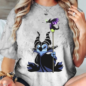May include: A gray t-shirt with a distressed look featuring a cartoon Stitch dressed as Maleficent from Disney's Sleeping Beauty. Stitch is holding a staff with a purple dragon on top.