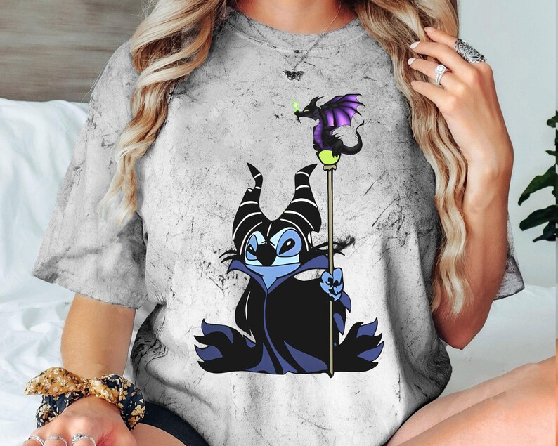 May include: A gray t-shirt with a distressed look featuring a cartoon Stitch dressed as Maleficent from Disney's Sleeping Beauty. Stitch is holding a staff with a purple dragon on top.