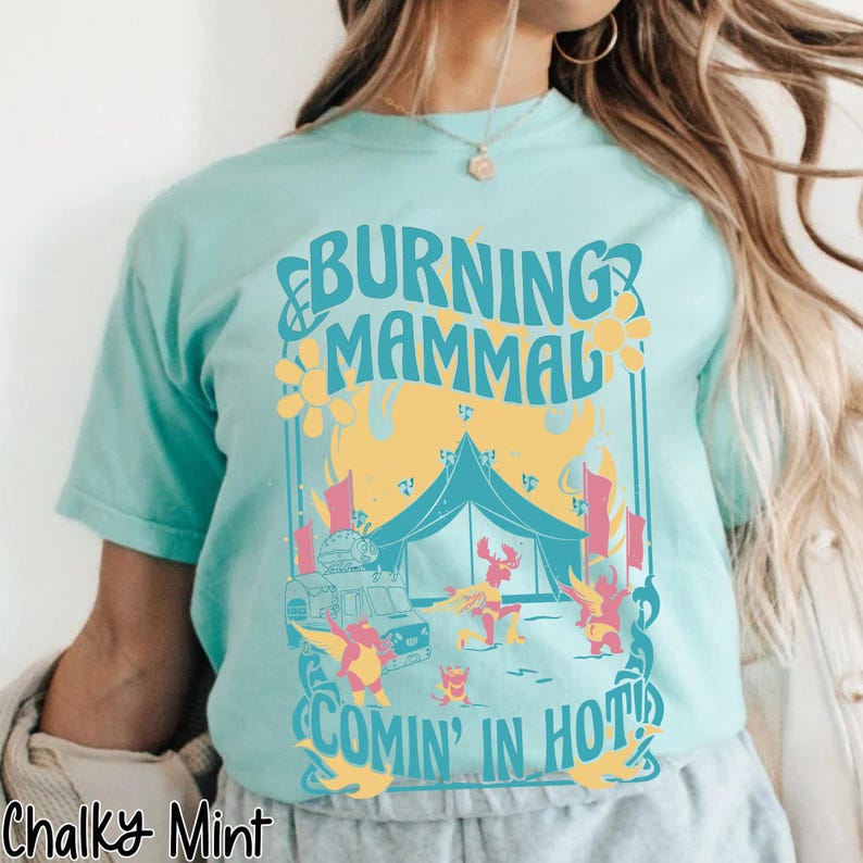 Puede incluir: Camiseta azul claro con un dise&ntilde;o gr&aacute;fico. El dise&ntilde;o presenta el texto "BURNING MAMMAL COMIN' IN HOT!". Muestra una tienda de campa&ntilde;a, una furgoneta y personajes de dibujos animados en una escena de festival. La camiseta es de un material suave y c&oacute;modo.