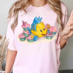 May include: Pink t-shirt featuring a cartoon illustration of a yellow fish with blue fins and a smiling face, surrounded by pink cupcakes with green frosting. The design is whimsical and colorful, perfect for fans of animated characters.