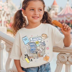 Puede incluir: Camiseta color crema con un dise&ntilde;o de dibujos animados para el D&iacute;a de los Profesionales Administrativos 2026. El gr&aacute;fico incluye una taza de caf&eacute;, una pila de papeles y un personaje de dibujos animados. La ni&ntilde;a sostiene un churro.
