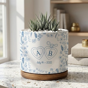 May include: A white ceramic planter with a blue floral design and a wooden base. The planter features two heart-shaped designs with the letters "A" and "B" and the date "July 11--2022". The planter holds a succulent plant.