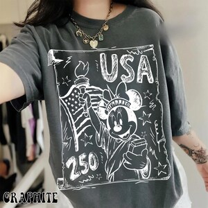 May include: Graphite-colored t-shirt featuring a white graphic of Minnie Mouse as the Statue of Liberty, holding a torch and a box, with the text "USA" and "250". The American flag is also depicted. The shirt has short sleeves.