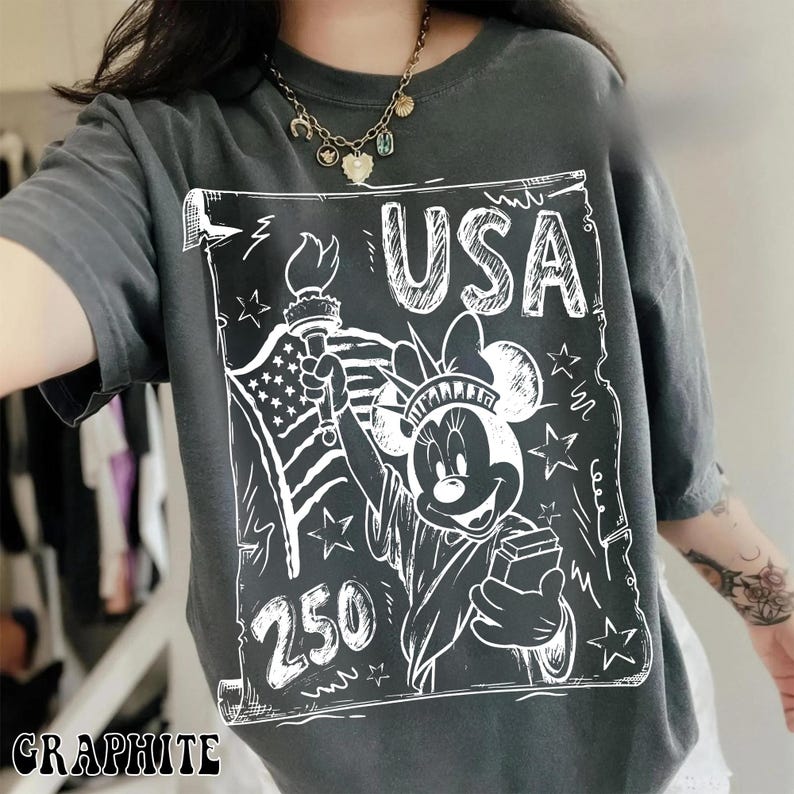 May include: Graphite-colored t-shirt featuring a white graphic of Minnie Mouse as the Statue of Liberty, holding a torch and a box, with the text "USA" and "250". The American flag is also depicted. The shirt has short sleeves.