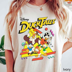 May include: A white t-shirt with a graphic print of the Disney DuckTales characters. The characters are in a retro 90s style with bright colors and geometric shapes. The text "Disney DuckTales" is printed in yellow letters.