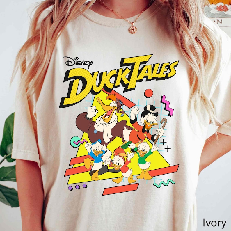 May include: A white t-shirt with a graphic print of the Disney DuckTales characters. The characters are in a retro 90s style with bright colors and geometric shapes. The text "Disney DuckTales" is printed in yellow letters.