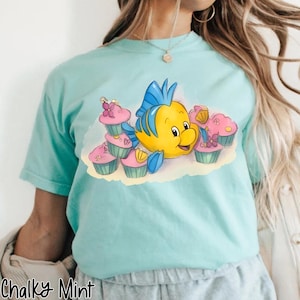 May include: A light blue t-shirt featuring a cartoon yellow fish with blue fins and a smiling face, surrounded by pink cupcakes with floral decorations. The text "Chalky Mint" is printed at the bottom left of the shirt.