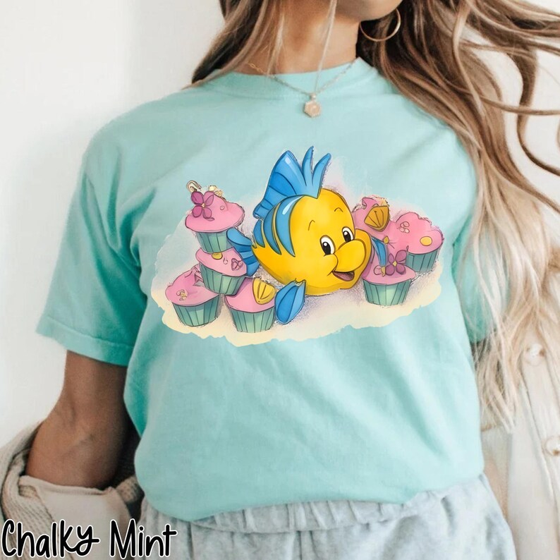 May include: A light blue t-shirt featuring a cartoon yellow fish with blue fins and a smiling face, surrounded by pink cupcakes with floral decorations. The text "Chalky Mint" is printed at the bottom left of the shirt.