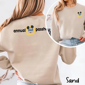 Puede incluir: Sudadera de color arena con las palabras "annual passholder" impresas en negro en la espalda. El dise&ntilde;o incluye un rat&oacute;n de dibujos animados con gafas de sol dentro de un cuadrado amarillo. Una versi&oacute;n m&aacute;s peque&ntilde;a del dise&ntilde;o est&aacute; en la parte delantera.