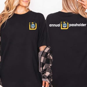 Puede incluir: Camiseta negra con un personaje de dibujos animados con gafas de sol dentro de un cuadrado amarillo. El texto "annual passholder" est&aacute; impreso en la espalda. Estilo casual.