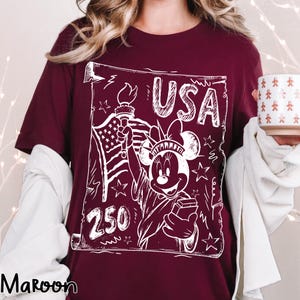 May include: Maroon t-shirt with a white graphic featuring the Statue of Liberty as Minnie Mouse, the American flag, and the word "USA". The number "250" is also included in the design. The shirt is a short-sleeved crew neck.