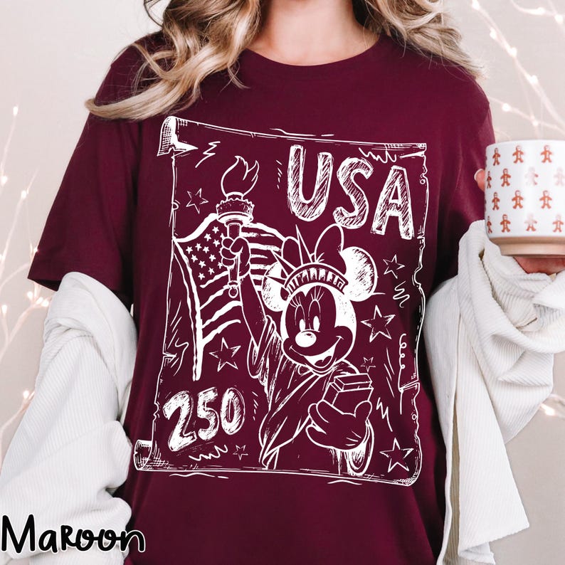 May include: Maroon t-shirt with a white graphic featuring the Statue of Liberty as Minnie Mouse, the American flag, and the word "USA". The number "250" is also included in the design. The shirt is a short-sleeved crew neck.