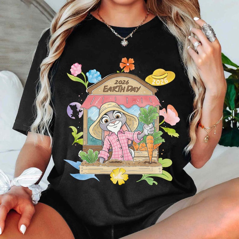 Op de afbeelding: Zwart T-shirt met een cartoonkonijn met een strohoed en een roze geruit overhemd, met wortels en groenten. De afbeelding bevat de tekst "2026 Earth Day" en bloemaccenten. Er is ook een gele hoed met "2026".