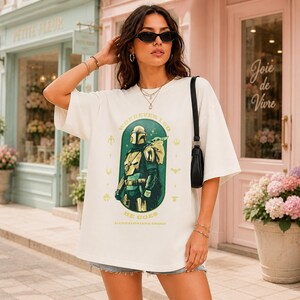 Peut inclure: T-shirt cr&egrave;me avec un motif de The Mandalorian et Grogu, avec le texte "WHEREVER I GO HE GOES". Le design est en vert, jaune et noir. Le t-shirt a des manches courtes et une coupe ample.