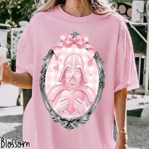 May include: Pink t-shirt featuring a framed illustration of Darth Vader holding a heart. The illustration is in shades of pink and has a gingham background with hearts. The word "Blossom" is printed at the bottom of the shirt.