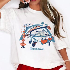 Op de afbeelding: Wit T-shirt met een kleurrijke afbeelding van een pretpark met een trein, raket en andere attracties. De woorden "LAND" en "Magic Kingdom" zijn ook zichtbaar in het ontwerp.