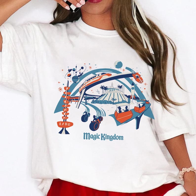 Op de afbeelding: Wit T-shirt met een kleurrijke afbeelding van een pretpark met een trein, raket en andere attracties. De woorden "LAND" en "Magic Kingdom" zijn ook zichtbaar in het ontwerp.