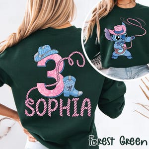 May include: Forest green sweatshirt with a pink and blue cowboy-themed design. Features a number 3, a cowboy hat, a boot, and the name "Sophia" in a rope-like font. The design also includes a cartoon character wearing a cowboy hat and holding a lasso.