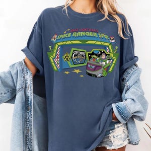 K&ouml;nnte beinhalten: Marineblaues T-Shirt mit einem Aufdruck von Buzz Lightyear's Space Ranger Spin. Das Design zeigt den Eingang der Attraktion, Buzz Lightyear und einen Alien-Charakter in einem Fahrzeug, mit dem Text "Buzz Lightyear Space Ranger Spin".