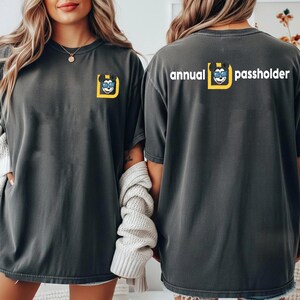 Puede incluir: Camisetas grises oscuras con un gr&aacute;fico de un personaje de dibujos animados en un cuadrado amarillo. El texto "annual passholder" est&aacute; impreso en blanco en la parte posterior. Las camisetas son de manga corta y de corte holgado.