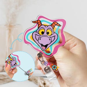 May include: A clear acrylic badge reel featuring a purple cartoon character with yellow eyes and orange horns, outlined with pink, blue, and yellow wavy lines. The reel has the word "EPCOT" in colorful letters. A smaller version of the character is shown in a circle.