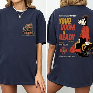 May include: Dark blue t-shirt with a hotel graphic and the text "Your Room is Ready" on the front. The back of the shirt features a cartoon bellhop and the text "We invite you if you dare, Your Room is Ready, Starring Your Bellhop, The Hollywood Tower Hotel."