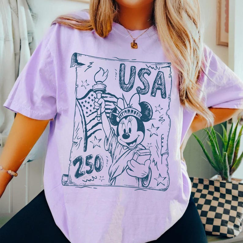 May include: A light purple t-shirt featuring a graphic of Minnie Mouse as the Statue of Liberty, holding a torch and a book, with the word "USA" above her. The American flag and the number "250" are also included in the design.