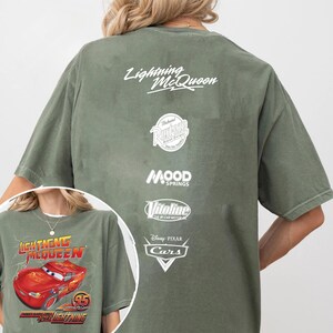 May include: Olive green t-shirt featuring Lightning McQueen from the Disney Pixar Cars movie. The front graphic shows Lightning McQueen with the text "Lightning McQueen" and "95". The back of the shirt has the text "Lightning McQueen" and various logos.
