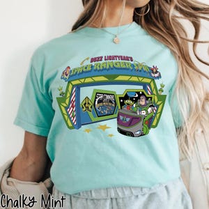 K&ouml;nnte beinhalten: Hellblaues T-Shirt mit einem Aufdruck von Buzz Lightyear's Space Ranger Spin. Das Design zeigt das Logo der Attraktion, Buzz Lightyear und andere Charaktere. Das Shirt ist unten mit "Chalky Mint" beschriftet.