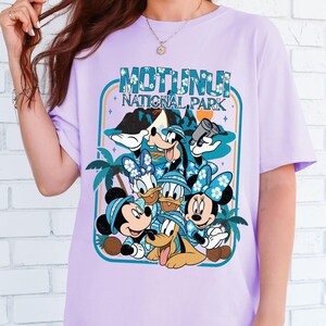 May include: A light purple t-shirt featuring a graphic of Disney characters, including Mickey, Minnie, Donald, Daisy, Goofy, and Pluto, with the text "MOTUNUI NATIONAL PARK" in a turquoise frame. The design includes palm trees and mountain scenery.