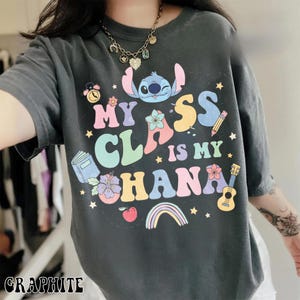 Puede incluir: Camiseta de color grafito con el texto "My Class is My Ohana" y un dibujo animado de Stitch. El dise&ntilde;o incluye un arco&iacute;ris, flores, un libro, un l&aacute;piz y un ukelele. La camiseta es de manga corta.